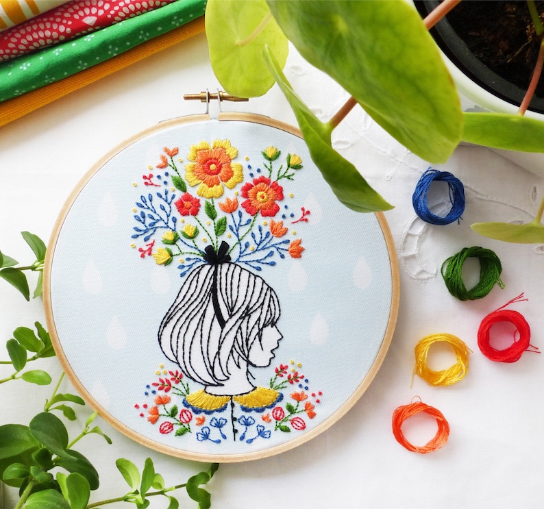 Op de afbeelding: Borduurwerk op een hoepel met een vrouw met een boeket kleurrijke bloemen op haar hoofd. De bloemen zijn in tinten rood, oranje, geel en groen. De vrouw heeft kort, zwart haar en draagt een gele kraag.