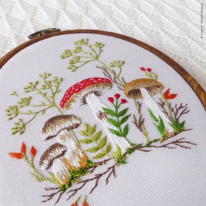 May include: Embroidered hoop art featuring a white background with three mushrooms, two brown and one red with white spots, surrounded by green foliage and red berries.