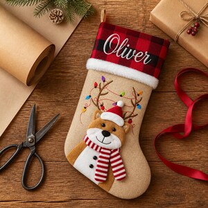 May include: A tan Christmas stocking featuring a red and black buffalo plaid cuff with the name Oliver embroidered in white script. The stocking displays a plush reindeer wearing a Santa hat and a red and white striped scarf with string lights.