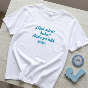 Könnte beinhalten: Ein weißes kurzärmeliges T-Shirt mit dem gestickten blauen Text "¿Qué mirás bobo? Anda pa'allá bobo" mittig auf der Brust. Das T-Shirt hat einen Rundhalsausschnitt und besteht aus schlichtem Stoff, präsentiert mit einer blauen sonnenförmigen Scheibe.