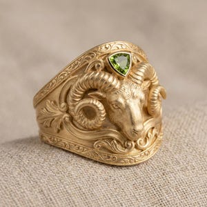 Może przedstawiać: Złoty pierścionek z misternym wzorem głowy barana. Pierścionek ma trójkątny zielony kamień szlachetny osadzony nad głową barana i jest ozdobiony skomplikowanymi ornamentami. Pierścionek jest prezentowany na teksturowanej, neutralnej powierzchni.