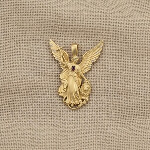 May include: A gold-colored pendant featuring an angel with outstretched wings, holding a cross and standing over a lion. The angel has a red gemstone in the center. The wings are adorned with small, clear stones. The pendant is on a tan background.