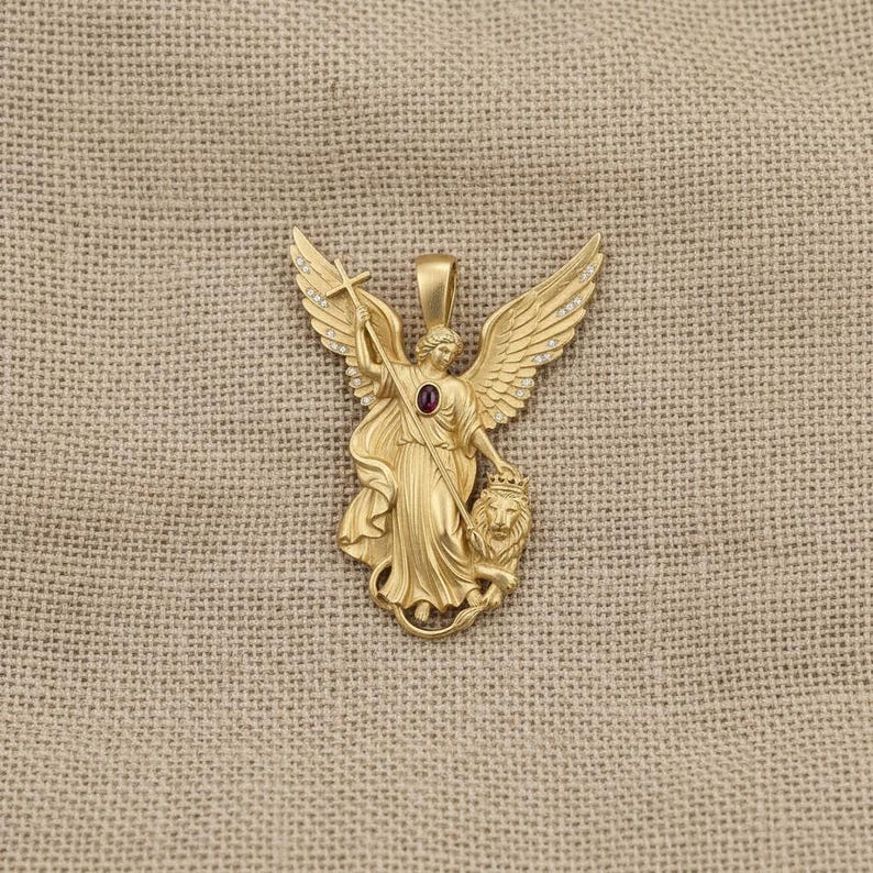 May include: A gold-colored pendant featuring an angel with outstretched wings, holding a cross and standing over a lion. The angel has a red gemstone in the center. The wings are adorned with small, clear stones. The pendant is on a tan background.