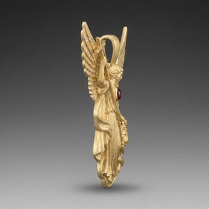 May include: A gold-colored angel pendant with outstretched wings, holding a cross. The pendant features a red gemstone and small, sparkling accents on the wings. The angel is depicted in flowing robes.