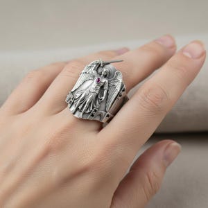 Pu&ograve; includere: Anello argentato con un dettagliato angelo che tiene una spada. L'angelo ha una gemma rossa al centro. L'anello, indossato su un dito, presenta un design dettagliato e in rilievo.