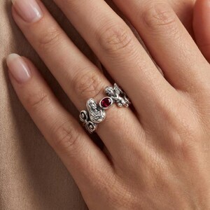 Puede incluir: Un anillo de plata con el perfil de una mujer, un peque&ntilde;o querub&iacute;n y una piedra preciosa roja. El anillo tiene un dise&ntilde;o ornamentado y detallado con una est&eacute;tica vintage. El anillo se lleva en un dedo.