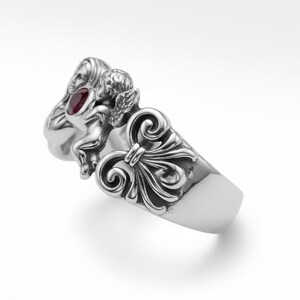 Puede incluir: Un anillo de plata con un dise&ntilde;o esculpido. Presenta un querub&iacute;n, un rostro femenino y adornos ornamentales. Una gema roja redonda est&aacute; engastada cerca del rostro de la mujer. El anillo tiene un acabado pulido y una banda ancha.