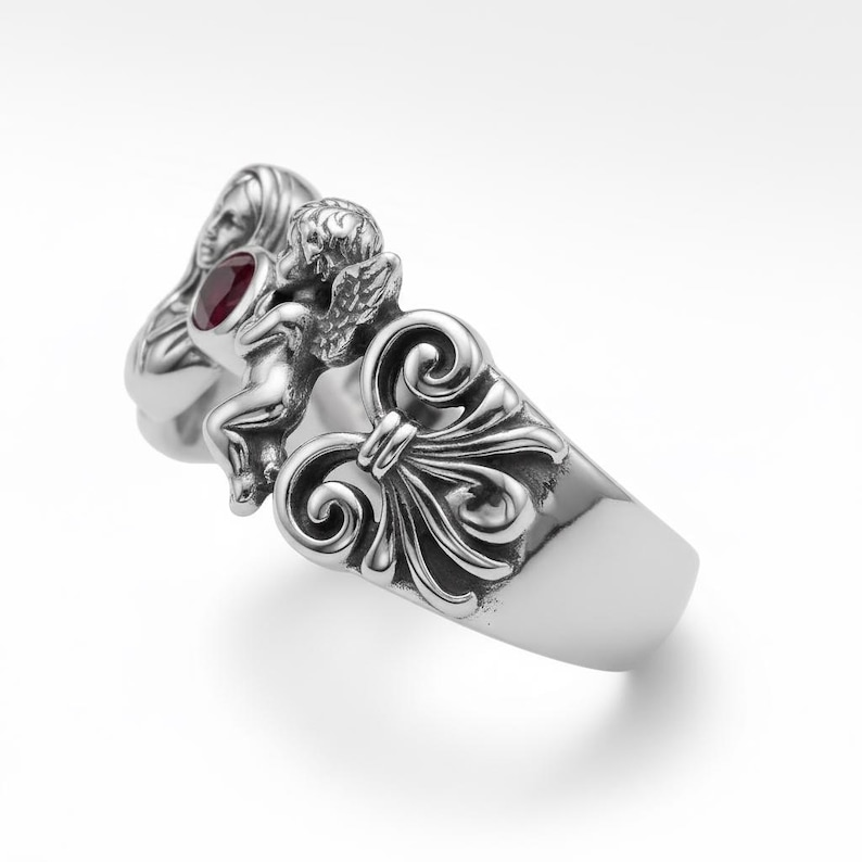 Puede incluir: Un anillo de plata con un dise&ntilde;o esculpido. Presenta un querub&iacute;n, un rostro femenino y adornos ornamentales. Una gema roja redonda est&aacute; engastada cerca del rostro de la mujer. El anillo tiene un acabado pulido y una banda ancha.