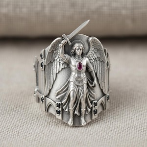 Pu&ograve; includere: Anello d'argento raffigurante un angelo con una spada alzata. L'angelo ha ali dettagliate e un'aureola. Una piccola gemma rossa ovale &egrave; incastonata nel petto. L'anello ha dettagli neri.
