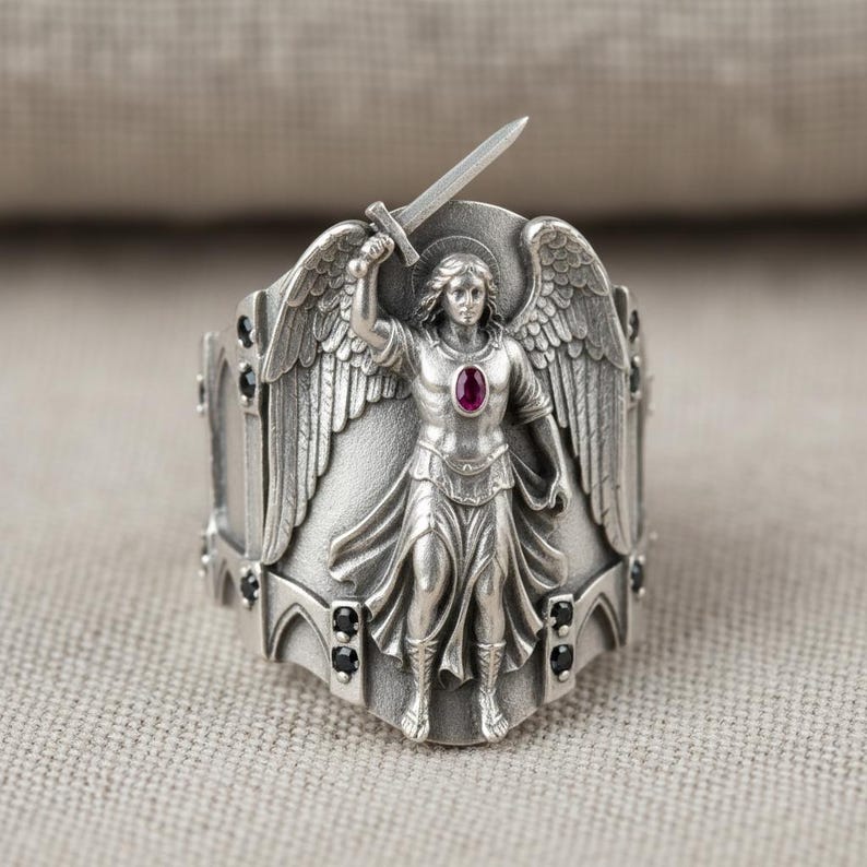 Pu&ograve; includere: Anello d'argento raffigurante un angelo con una spada alzata. L'angelo ha ali dettagliate e un'aureola. Una piccola gemma rossa ovale &egrave; incastonata nel petto. L'anello ha dettagli neri.
