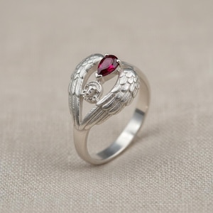 Könnte beinhalten: Ein silberner Ring mit Engelsflügel-Design und einem birnenförmigen, tiefroten Edelstein. Der Ring weist detaillierte Federstrukturen und ein kleines, kreisförmiges Emblem auf. Der Ring ist vor einem neutralen Hintergrund platziert.