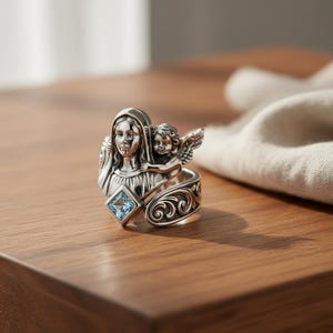 May include: A silver ring featuring a religious theme. The ring showcases a sculpted figure of a woman with a child angel, and a square, light blue gemstone. The band has an ornate, swirling design.