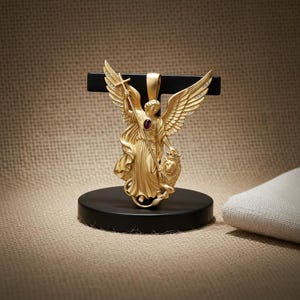 May include: A gold-colored pendant featuring an angel with outstretched wings, holding a cross and a lion. The angel has a red gemstone in the center. The pendant is displayed on a black stand.