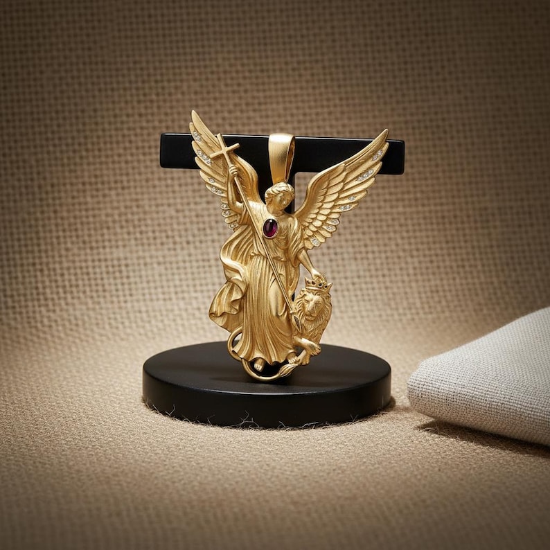 May include: A gold-colored pendant featuring an angel with outstretched wings, holding a cross and a lion. The angel has a red gemstone in the center. The pendant is displayed on a black stand.
