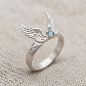 May include: A silver ring featuring a detailed wing design with a small, round, light blue gemstone. The ring has a deer head engraving on the band. The wings are positioned to appear as if they are in flight, creating a unique and artistic jewelry piece.