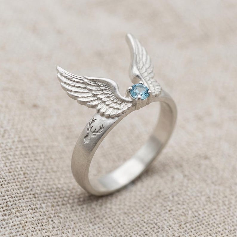 May include: A silver ring featuring a detailed wing design with a small, round, light blue gemstone. The ring has a deer head engraving on the band. The wings are positioned to appear as if they are in flight, creating a unique and artistic jewelry piece.
