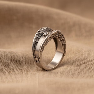 May include: A silver ring with detailed angel wing designs. The ring features a small, clear gemstone at the centre and a decorative emblem on the band. The wings are intricately carved, showcasing a feathered texture.