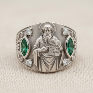 May include: A silver ring featuring a detailed relief of a bearded figure holding tablets with Hebrew text. The ring is adorned with emerald-green and clear gemstones, set in an ornate design. The ring has a polished finish.