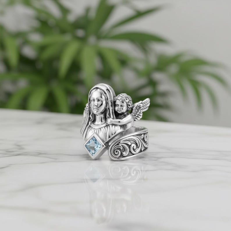 May include: A silver ring featuring a sculpted design of a woman and an angel. The ring has a square, light blue gemstone and ornate scrollwork. The ring is a religious-themed jewelry item.