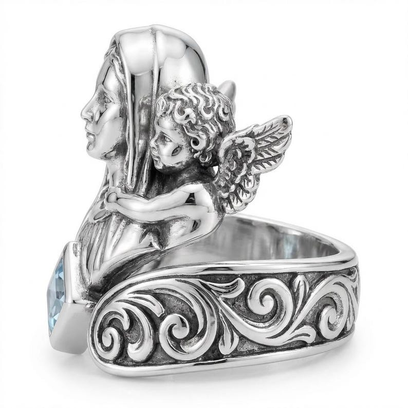 May include: A silver ring featuring a sculpted design of a woman and an angel. The ring has a light blue gemstone and a band with an ornate, swirling pattern. The angel has detailed wings, and the woman is wearing a head covering.