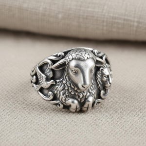 May include: A silver ring with a detailed sheep design at the centre, accompanied by a dove and a lion. The ring has an ornate, textured appearance with intricate detailing. A statement piece of jewellery.