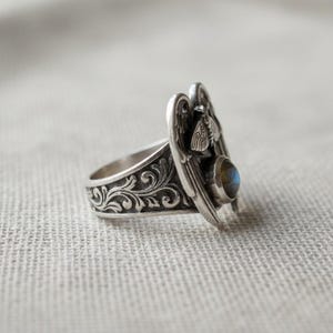 May include: A silver ring featuring an ornate design with a central moth and a small, round, blue-grey gemstone. The ring has detailed scrollwork on the band and wings on the top.