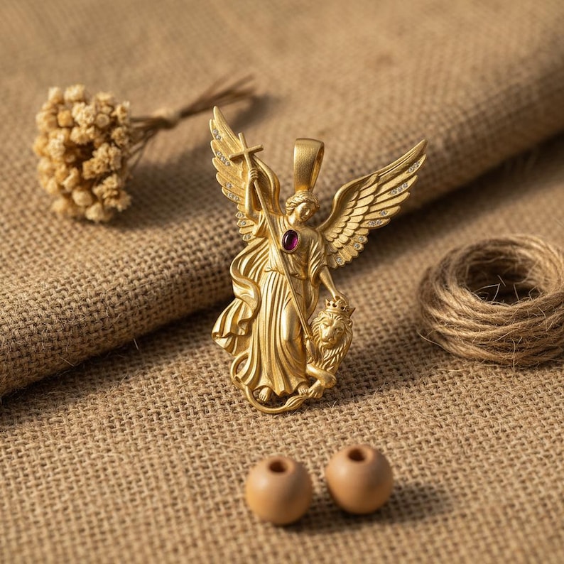 May include: A gold-colored pendant featuring an angel with outstretched wings, holding a cross and standing over a lion. The pendant has a red gemstone in the center. The image also includes dried flowers, twine, and two wooden beads.