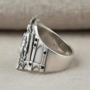 Pu&ograve; includere: Un anello d'argento con un rilievo dettagliato di una figura in un abito lungo, una piccola pietra rossa e dettagli neri. L'anello ha una fascia larga e un design architettonico sul lato.