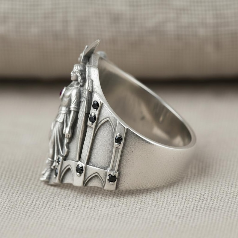 Pu&ograve; includere: Un anello d'argento con un rilievo dettagliato di una figura in un abito lungo, una piccola pietra rossa e dettagli neri. L'anello ha una fascia larga e un design architettonico sul lato.