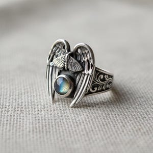 May include: A silver ring featuring an angel wing design with a central, iridescent, round gemstone. The ring has a detailed, ornate pattern on the band and a small moth figure between the wings.