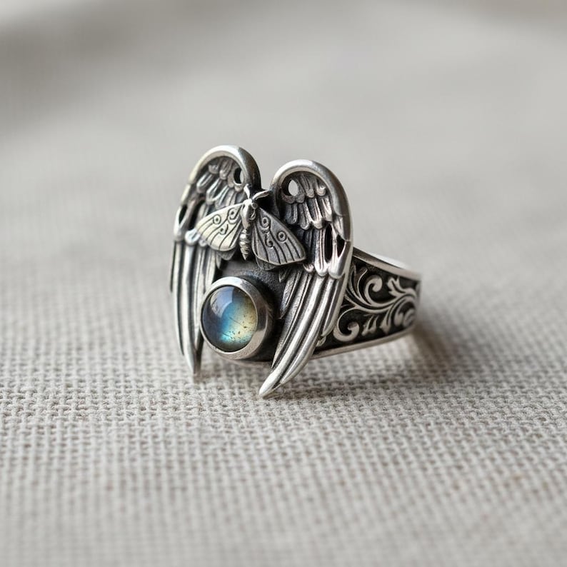 May include: A silver ring featuring an angel wing design with a central, iridescent, round gemstone. The ring has a detailed, ornate pattern on the band and a small moth figure between the wings.