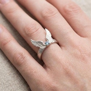 May include: A silver ring featuring a detailed wing design with a small, round, light blue gemstone at the center. The ring's wings are spread open, creating a unique and elegant look. The ring is shown on a person's finger.