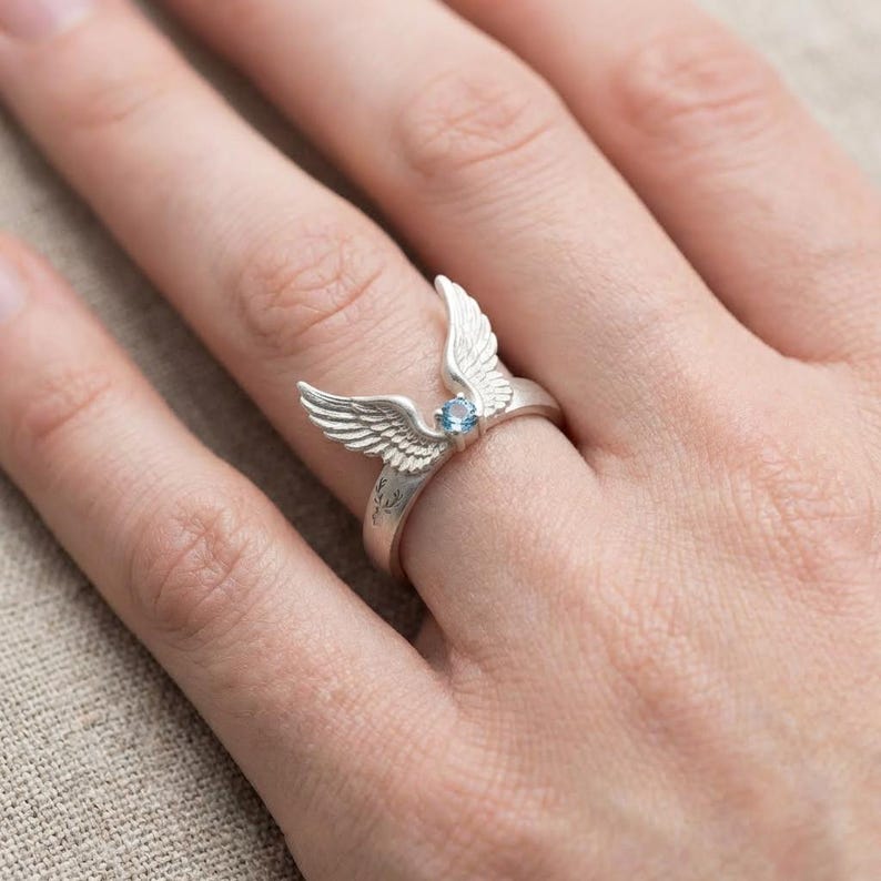 May include: A silver ring featuring a detailed wing design with a small, round, light blue gemstone at the center. The ring's wings are spread open, creating a unique and elegant look. The ring is shown on a person's finger.