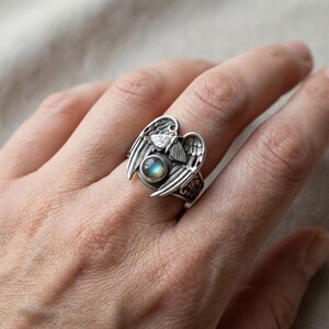 May include: A silver ring featuring an angel wing design. The ring has a central, round, blue-green stone and intricate details. The wings are spread out on either side of the stone, with a small butterfly design above the stone.