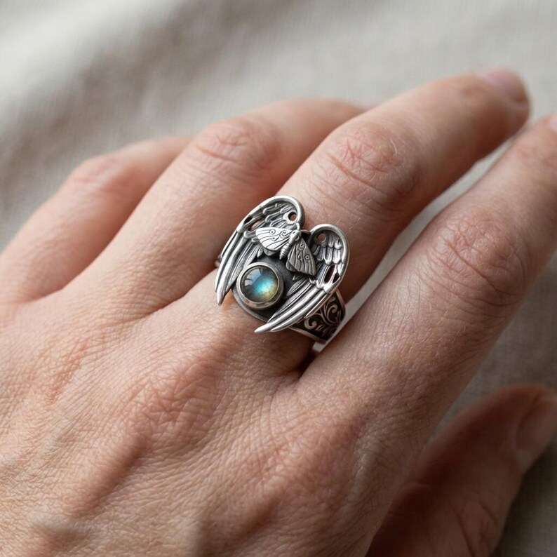 May include: A silver ring featuring an angel wing design. The ring has a central, round, blue-green stone and intricate details. The wings are spread out on either side of the stone, with a small butterfly design above the stone.
