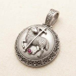 Puede incluir: Colgante de plata con un relieve detallado de un cordero sosteniendo una cruz, dentro de un marco circular. El cordero tiene detalles de lana rizada y una pequeña gema roja. El colgante tiene un asa adornada con un diseño de clave griega.
