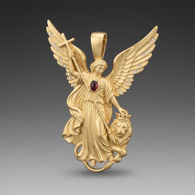 May include: A gold-colored pendant featuring an angel with outstretched wings, holding a cross and standing beside a crowned lion. The angel's dress has a red gemstone. The wings are adorned with small, clear stones. The pendant has a loop for a chain.