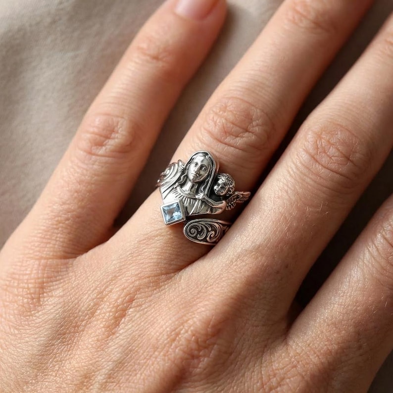 May include: A silver-toned ring featuring a detailed design of a woman and a cherub, with a small, square, light blue gemstone. The ring has an ornate, swirling pattern and is worn on a finger.