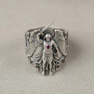 Pu&ograve; includere: Anello d'argento raffigurante un angelo con ali e spada. L'angelo ha un'aureola e una gemma rossa. L'anello presenta elementi decorativi con accenti neri. Gioiello dettagliato.