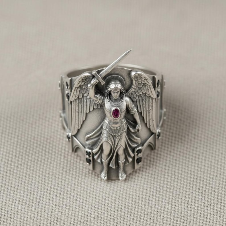 Pu&ograve; includere: Anello d'argento raffigurante un angelo con ali e spada. L'angelo ha un'aureola e una gemma rossa. L'anello presenta elementi decorativi con accenti neri. Gioiello dettagliato.