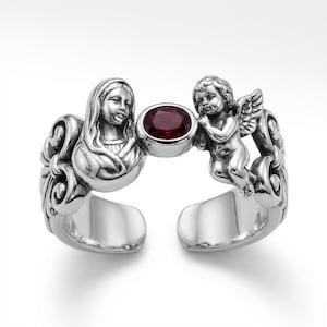 Puede incluir: Anillo plateado con un dise&ntilde;o esculpido que presenta un busto de mujer y un querub&iacute;n alado. Una gema ovalada de color rojo oscuro se encuentra entre las figuras. El anillo tiene un dise&ntilde;o abierto.