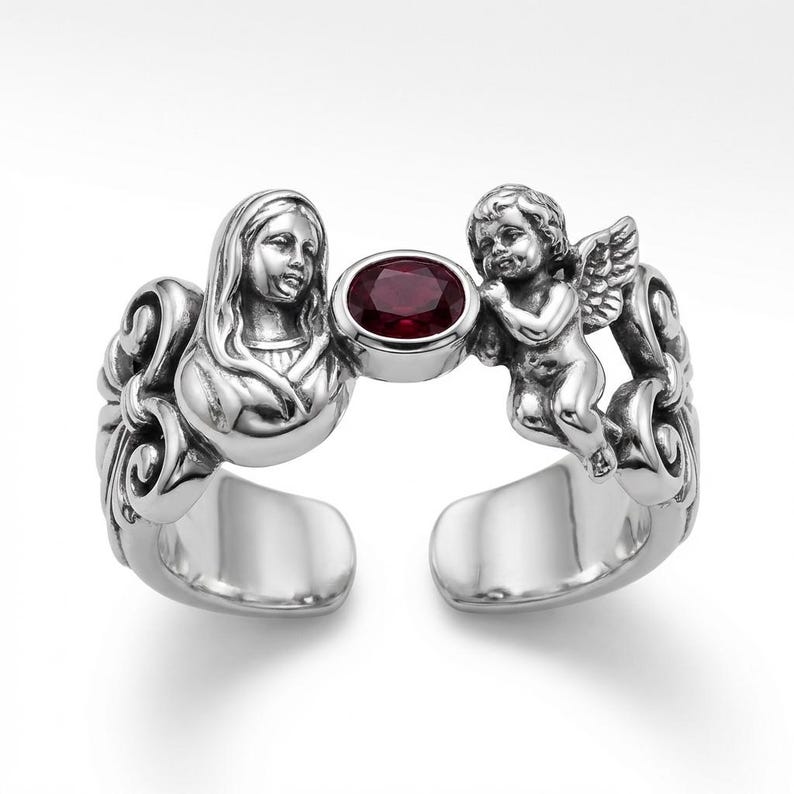 Puede incluir: Anillo plateado con un dise&ntilde;o esculpido que presenta un busto de mujer y un querub&iacute;n alado. Una gema ovalada de color rojo oscuro se encuentra entre las figuras. El anillo tiene un dise&ntilde;o abierto.