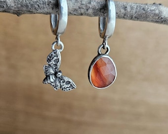 May include: A pair of silver hoop earrings with a dangling charm. One earring has a silver butterfly charm, and the other has a teardrop-shaped orange carnelian stone.