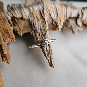 May include: A silver ring with a small, oval-shaped design featuring a sunburst pattern.