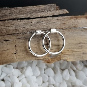 May include: A pair of silver hoop earrings with a slightly hammered texture. The earrings are on a wooden surface with white stones.