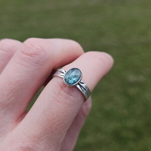 Puede incluir: Un anillo de plata con una piedra preciosa ovalada de color turquesa. El anillo tiene un diseño de banda simple y moderno. La piedra preciosa está engastada en un bisel. El anillo se muestra en un dedo sobre un fondo verde borroso.