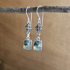 May include: Silver dangle earrings with square, light green gemstones. The earrings feature a geometric design with a starburst pattern above the gemstone. The earrings are hanging from a small, brown branch.