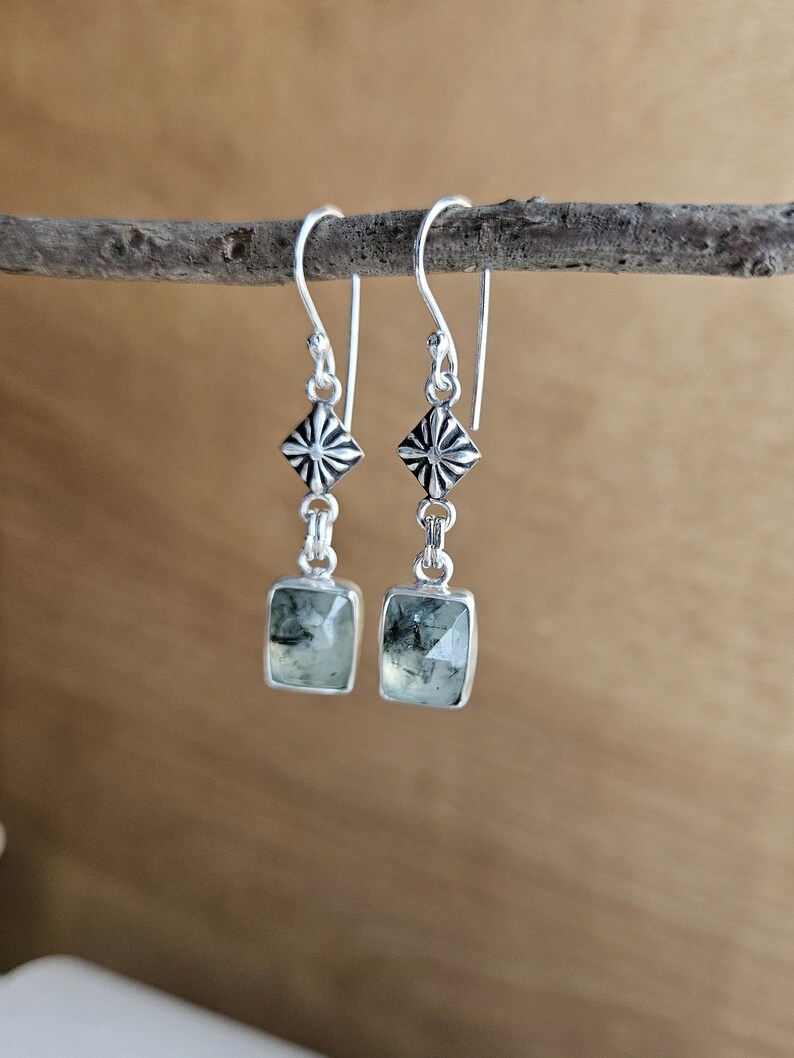 May include: Silver dangle earrings with square, light green gemstones. The earrings feature a geometric design with a starburst pattern above the gemstone. The earrings are hanging from a small, brown branch.