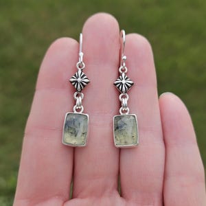 May include: A pair of silver earrings with a rectangular, light green stone. The earrings have a decorative, diamond-shaped element above the stone. The background is a blurred green.