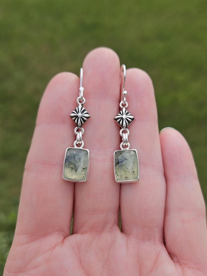 May include: A pair of silver earrings with a rectangular, light green stone. The earrings have a decorative, diamond-shaped element above the stone. The background is a blurred green.
